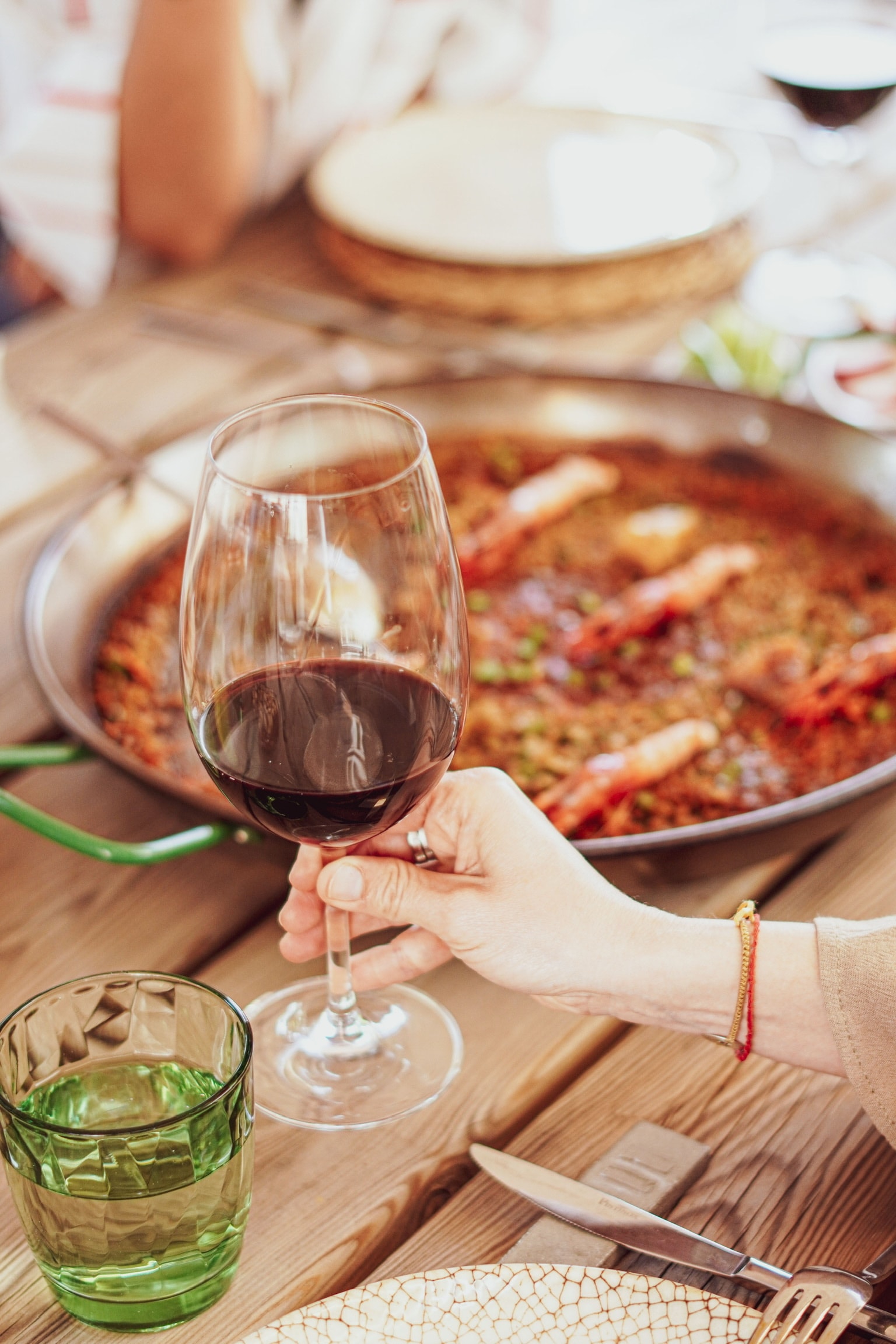 Una mà alça una copa de vi negre damunt una paella d'arròs a taula
